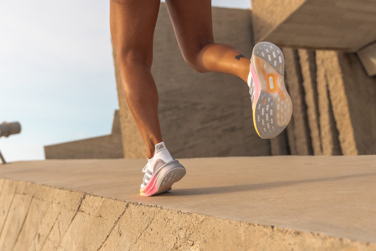 Low-angle close-up of Kaja Časar’s legs mid-stride, showing the sculpted outsole of the Adidas Ultraboost Light with its pink‑to‑yellow Lightboost gradient, captured against warm-toned brutalist architecture at sunrise.